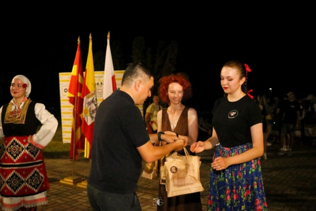 Tancerka w polskim stroju ludowym z opiekunką odbiera od mężczyzny podarunki , w tle flagi Macedonii i Polski