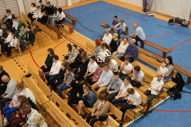 Na sali gimnastycznej siedzą rodzice, uczniowie i nauczyciele podczas Święta KEN w szkole w Niebocku