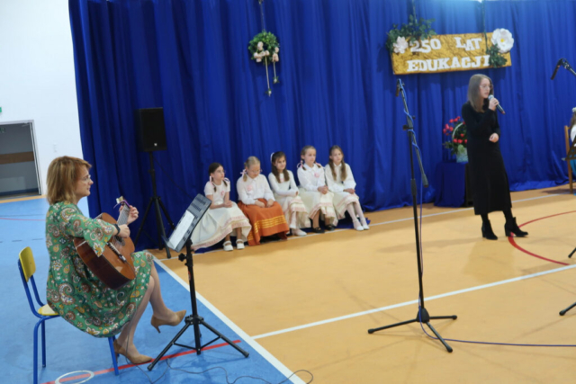 Uczennica klasy VIII podczas wykonania piosenki "Pokolenie" do akompaniamentu gitary. W tle dekoracja oraz uczennice klas młodszych.