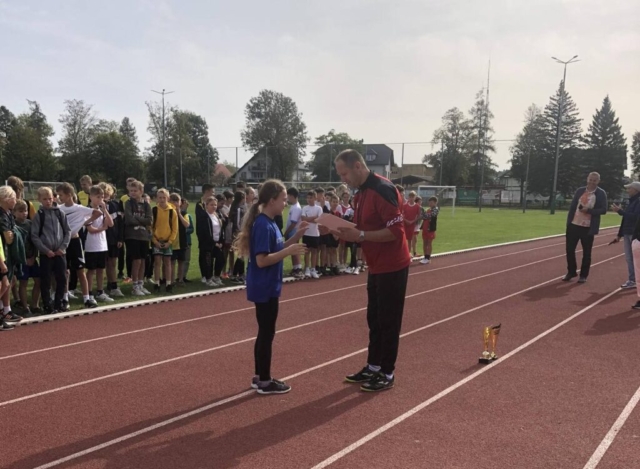 Dziewczynka, zawodniczka reprezentująca szkołę w Niebocku odbiera dyplom na zawodach sportowych, w tle stadion sportowy i inni zawodnicy
