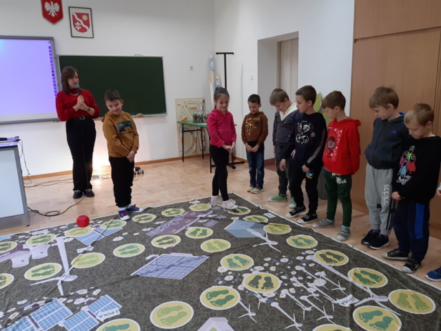 Grupa dzieci stoi przed rozłożoną na podłodze planszą do gry. Dzieci odpowiadają na pytania prowadzącej.