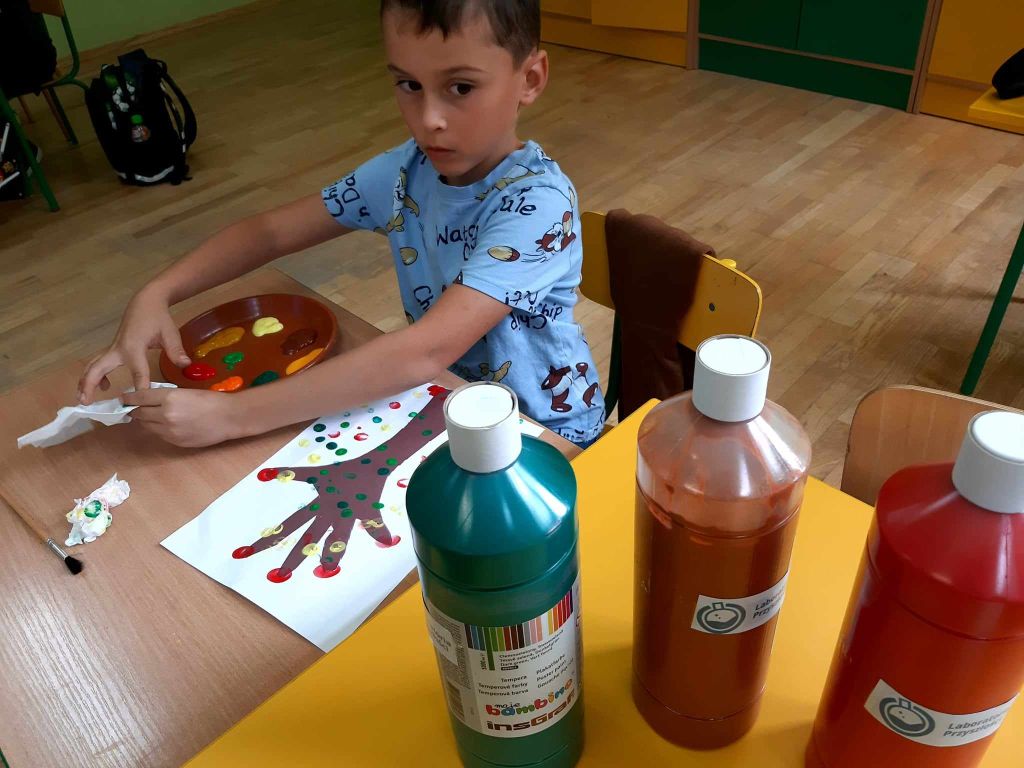 Uczeń wykonuje pracę plastyczną za pomocą palców i farb, na stoliku stoją butelki z farbą i talerz papierowy służący za paletę malarską