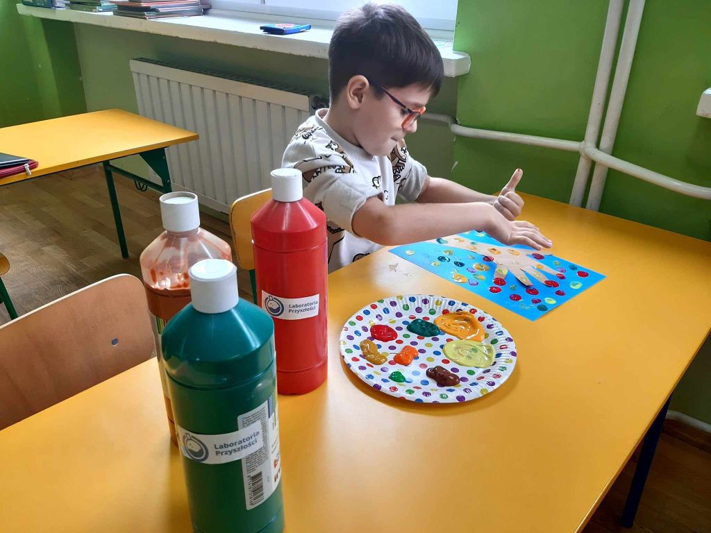 Uczeń wykonuje pracę plastyczną za pomocą palców i farb, na stoliku stoją butelki z farbą i talerz papierowy służący za paletę malarską