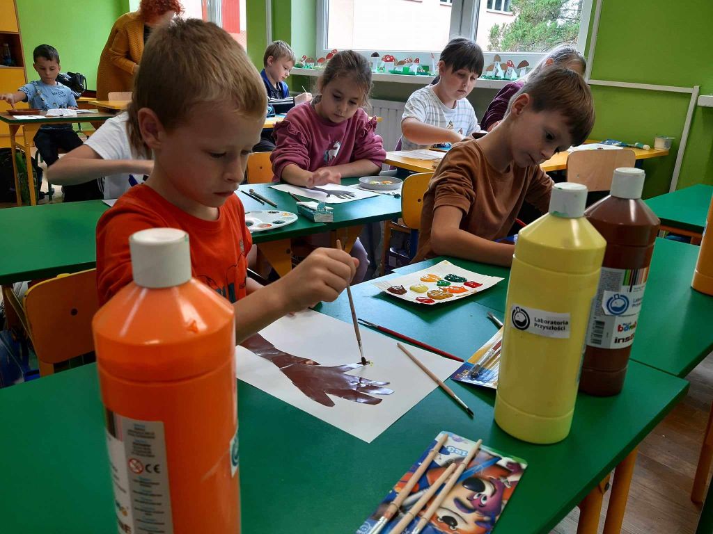 Uczniowie siedzą w ławkach i malują farbami
