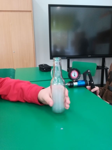Na stole stoi butelka trzymana przez osobę w czerwonej bluzie, wewnątrz butelki widoczna jest mgła.