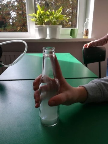 Na zielonym stoliku stoi butelka wypełniona mgłą wywołaną doświadczeniem z rozprężaniem powietrza.