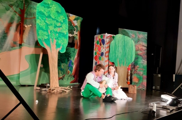 Na scenie dwoje aktorów siedzi na podłodze i rozmawia ze sobą. Dekoracja w tle przedstawia las.