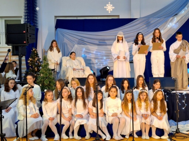 Na tle świątecznej dekoracji dzieci w białych strojach odgrywają scenki jasełkowe
