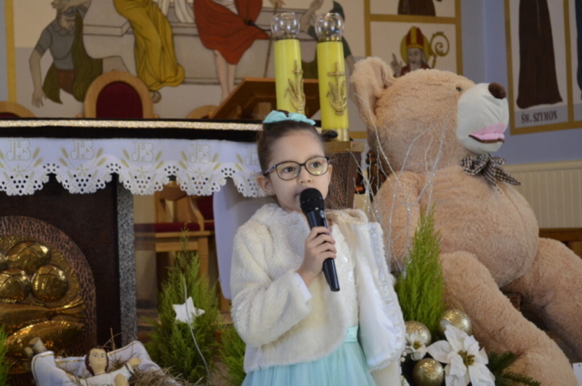 Dziewczynka stoi z mikrofonem i śpiewa kolędę, w tle wnętrze kościoła i maskotka miś jako nagroda