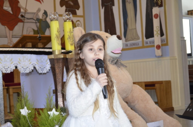 Dziewczynka stoi z mikrofonem i śpiewa kolędę, w tle wnętrze kościoła i maskotka miś jako nagroda