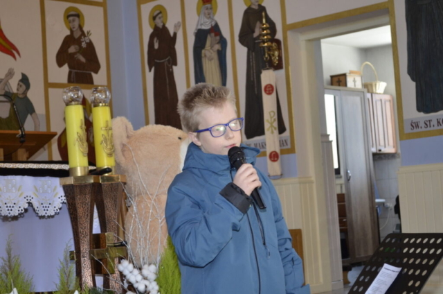 Chłopiec stoi z mikrofonem i śpiewa kolędę, w tle wnętrze kościoła i maskotka miś jako nagroda