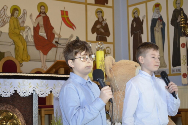 Dwaj chłopcy stoją z mikrofonami i śpiewają kolędę, w tle wnętrze kościoła i maskotka miś jako nagroda
