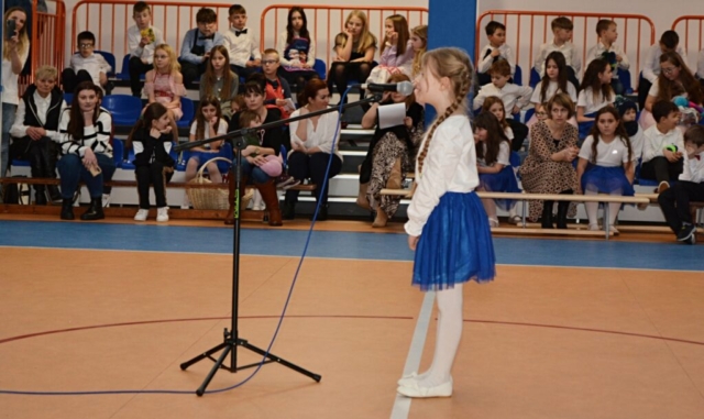 Dziewczynka stoi przed mikrofonem i mówi wierszyk. w tle publiczność, dzieci, rodzice, nauczyciele