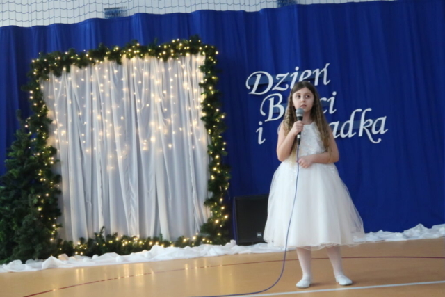 Dziewczynka w białej sukience trzyma mikrofon i śpiewa piosenkę, w tle dekoracja okolicznościowa