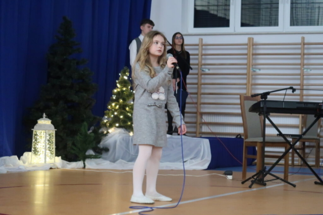 Dziewczynka w szarej sukience trzyma mikrofon i śpiewa piosenkę, w tle dekoracja okolicznościowa oraz para konferansjerów