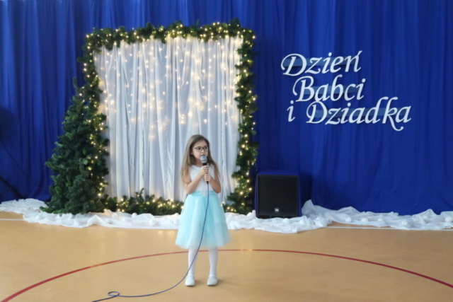 Dziewczynka na tle dekoracji okolicznościowej z napisem Dzień Babci i Dziadka stoi z mikrofonem i śpiewa