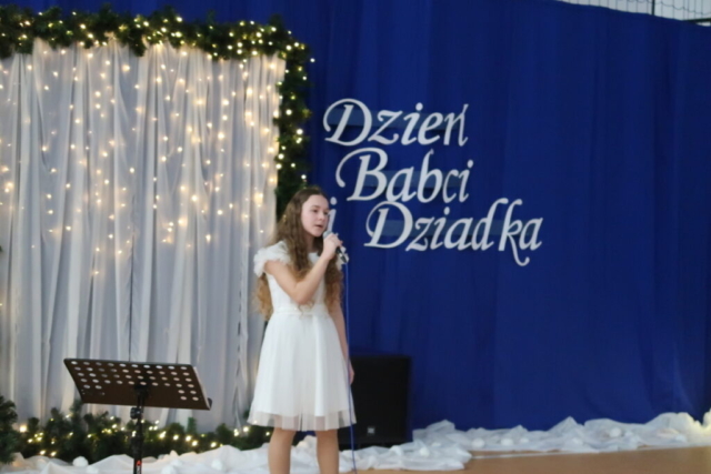 Dziewczynka w białej sukience trzyma mikrofon i śpiewa piosenkę, w tle dekoracja okolicznościowa