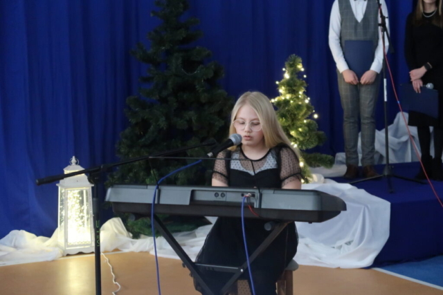 Dziewczynka gra na keyboardzie i śpiewa do mikrofonu, w tle granatowa kurtyna i elementy dekoracji świątecznej