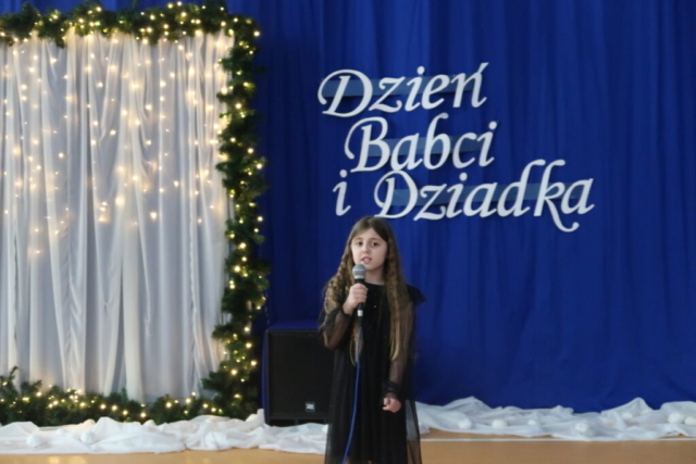 Dziewczynka w czarnej sukience trzyma mikrofon i śpiewa piosenkę, w tle dekoracja okolicznościowa