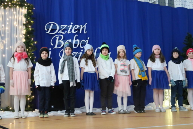 Grupa dzieci z klasy II w czapeczkach, szalikach i rękawiczkach stoi na tle dekoracji i występuje z piosenką i tańcem