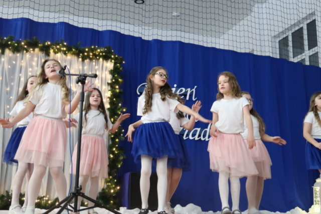 Dziewczynki w białych koszulkach, różowych i niebieskich spódniczkach tańczą sambę i śpiewają piosenkę, na drugim planie dekoracja okolicznościowa