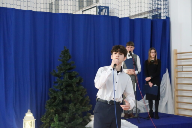 Uczeń stoi z mikrofonem na tle dekoracji i wykonuje piosenkę, Z tyłu para konferansjerów