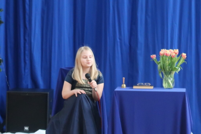 Dziewczynka odgrywa główną rolę hrabiny, siedzi przy stole w eleganckiej sukni, obok wazon z kwiatami, w tle granatowa kurtyna