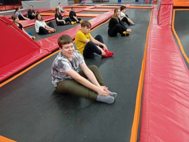 Dziewczynki i chłopcy wykonują ćwiczenia rozgrzewające siedząc na trampolinach