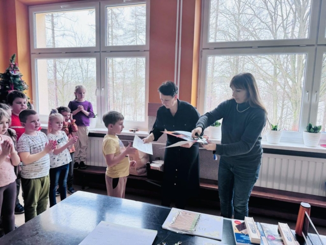 Chłopiec w żółtej koszulce z rąk pani dyrektor otrzymuje dyplom i nagrodę, w tle grupa uczniów stojąca na korytarzu szkolnym