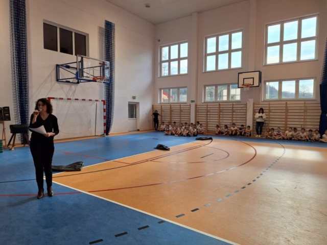 nauczycielka na sali gimnastycznej ogłasza rozpoczęcie rozgrywek. W tle dzieci ubrane w stroje sportowe.