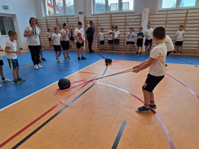 Dzieci w strojach sportowych biorą udział w rywalizacjach - ciągnięcie piłki w hula-hop