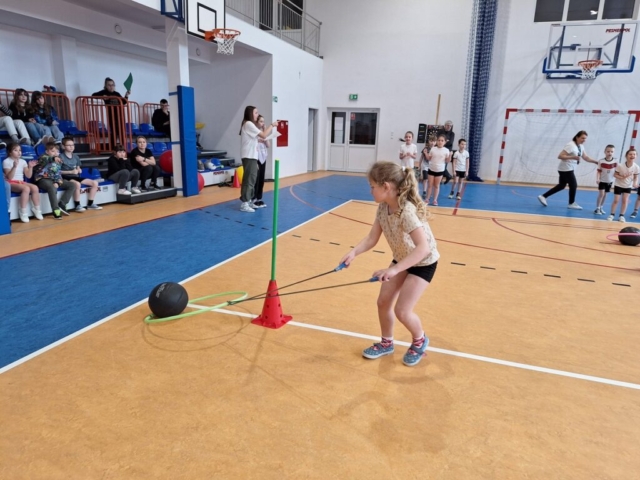 Dzieci w strojach sportowych biorą udział w rywalizacjach - ciągnięcie piłki w hula-hop