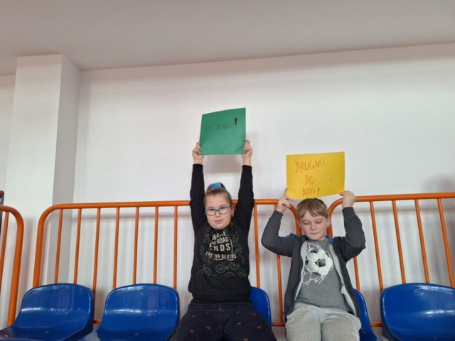 Dwoje dzieci siedzi na trybunach, w górze trzyma plakaty dopingujące kolegów w rozgrywkach