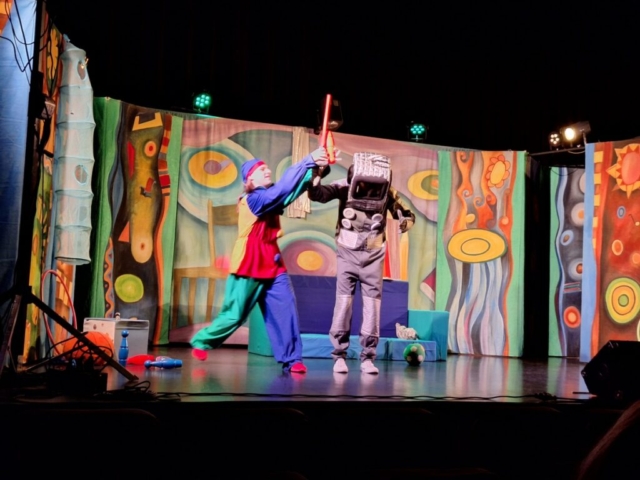 Dwoje aktorów na scenie odgrywa rolę pajacyka i robota. W tle barwna dekoracja przedstawiająca dziecięcy pokój