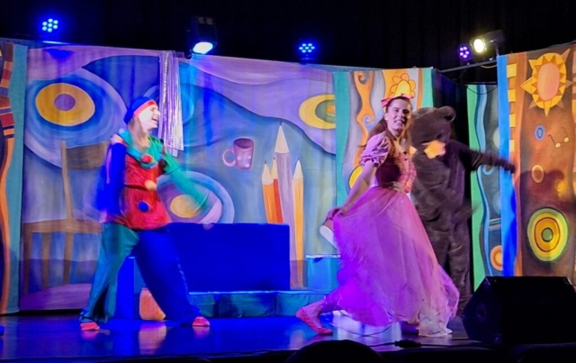 Troje aktorów - lalka, miś i pajac tańczą i śpiewają na scenie. W tle barwna dekoracja przedstawiająca dziecięcy pokój