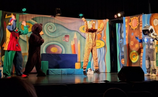 Czworo aktorów - tygrysek, miś, robot i pajac odgrywają scenkę teatralną. W tle barwna dekoracja przedstawiająca dziecięcy pokój