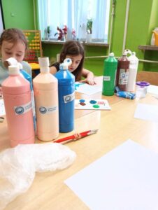 Dwie dziewczynki siedzą przy stoliku i pracują nad pracą plastyczną wykorzystując farby 