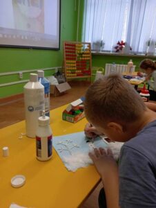 Chłopiec siedzi przy stoliku w klasie i maluje pracę plastyczną z wykorzystaniem farb, wycinanek i waty