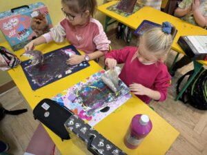 Dziewczynki z klasy 1 malują farbkami prace plastyczne siedząc w ławeczkach szkolnych