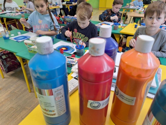Butelki farby stojące na stoliku, w tle uczniowie wykonujący prace plastyczne.