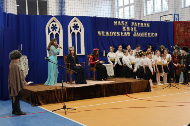 Dzieci szkolne przedstawiają scenkę nawiązującą do życia królowej Jadwigi i Władysława Jagiełły. Obok sceny zgromadzony jest szkolny chórek