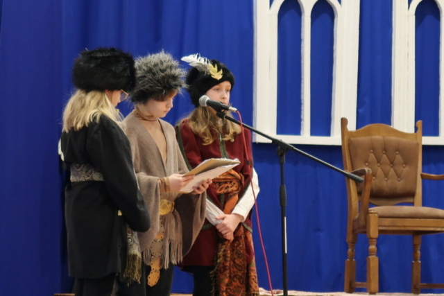 Troje dzieci w strojach średniowiecznych odgrywają na tle dekoracji scenkę nawiązującą do życia Patrona szkoły