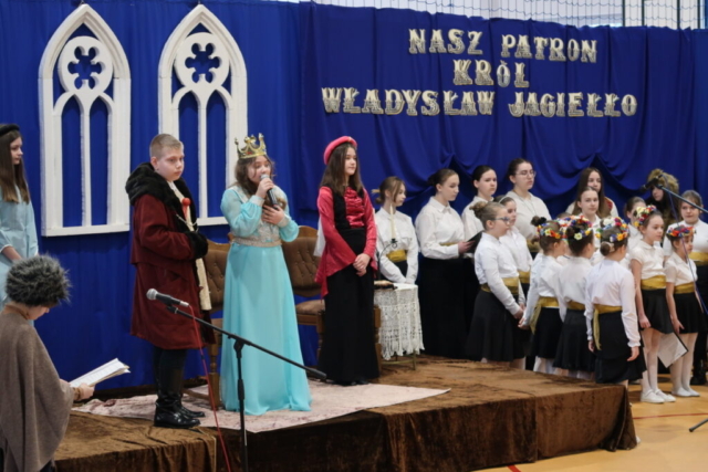 Dzieci w strojach średniowiecznych odgrywają na tle dekoracji scenkę nawiązującą do życia Patrona szkoły i królowej Jadwigi. Na drugim planie szkolny chórek.
