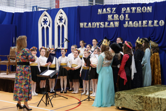 Chór szkolny wykonuje utwór "Gaude Mater Polonia". Nauczyciel dyryguje śpiewem. W tle dekoracja związana z obchodzonym świętem