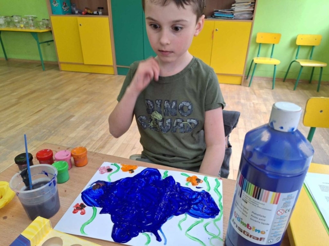 Chłopiec siedzi w ławce szkolnej i wykonuje pracę plastyczną za pomocą farb