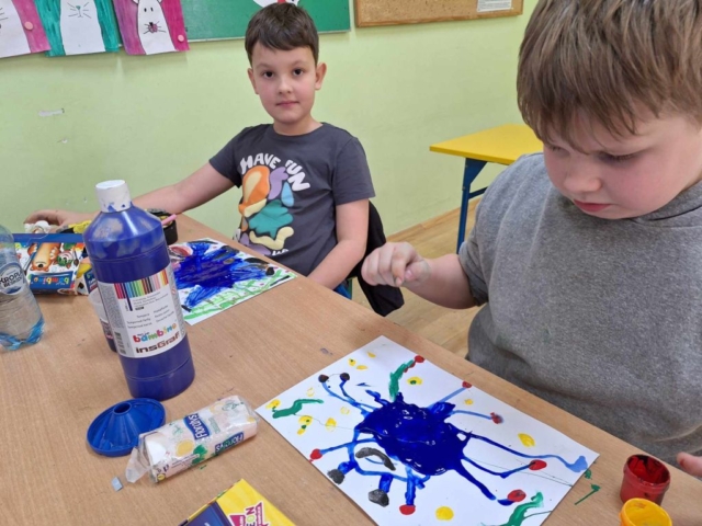 Dwóch chłopców siedzi w ławce szkolnej i wykonuje prace plastyczne za pomocą farb